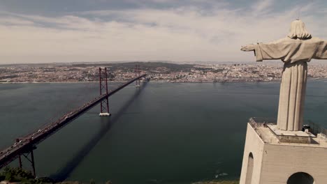 Aerial-pull-out-shot-overlooking-at-Lisbon-capital-city,-reveals-clifftop-statue-of-Cristo-Rei-in-Almada