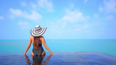 Woman-in-black-bathing-suit-and-large-hat-sitting-on-edge-of-infinity-swimming-pool-overlooking-turquoise-ocean-raises-arms-while-enjoying-view-of-stunning-beachside-tropical-resort-destination