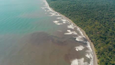 Aerial-shot-of-dirty-brown-coastline-of-Costa-Rica-beside-green-lush-forest-trees