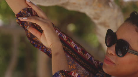 Close-up-of-Latina-Girl-Model-Wearing-Sunglasses-Poses-next-to-Beachside-Tropical-Tree,-tilt-reveal-shot