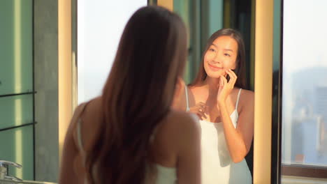 Woman-checking-and-touching-her-face-skin-near-the-mirror-smiling,-over-the-shoulder-shot