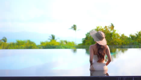 Vista-Trasera-De-La-Mujer-Sentada-Al-Borde-De-La-Piscina-Infinita-En-Un-Hotel-Exótico-En-Bali-Usando-Monokini-Y-Sombrero-Para-El-Sol