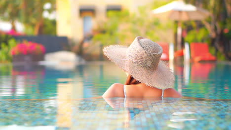 Back-view-Portrait-of-an-unrecognizable-woman-inside-swimming-pool-water-in-a-sunhat,-blurred-tropical-lounge-on-background