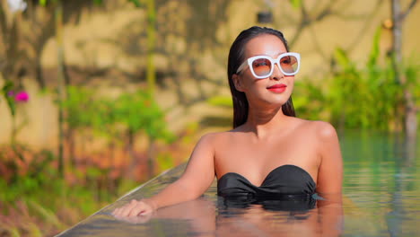 Portrait-of-a-happy-smiling-brunette-woman-in-black-swimsuit-relaxing-inside-swimming-pool-and-looking-aside-in-Thailand-resort