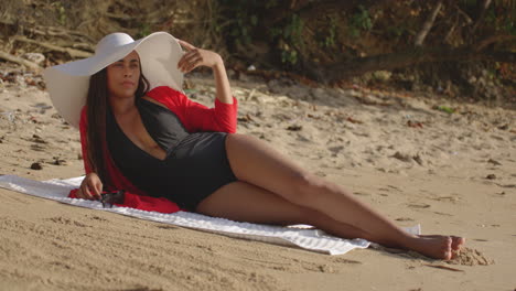 Lateinische-Modellfrau-Am-Strand-Mit-Hut,-Die-Sich-Auf-Die-Seite-Legt,-Setzt-Eine-Sonnenbrille-Mit-Blick-Auf-Die-Kamera-Auf