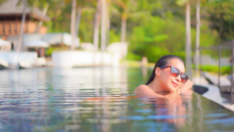 Beautiful-young-Asian-woman-wearing-sunglasses-resting-at-side-of-infinity-swimming-pool-of-luxury-tropical-island-resort-and-spa