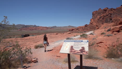 Back-of-Young-Female-Landscape-Photographer-in-Valley-of-Fire-State-Park,-Nevada-USA-Passing-by-Balancing-Rock-Trailhead-Sign,-Slow-Motion