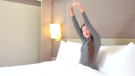 Pretty-Asian-woman-with-long-hair-wearing-grey-top-stretches-arms-over-head-with-eyes-closed-sitting-in-bed-with-comfortable-puffy-white-linen-and-pillows