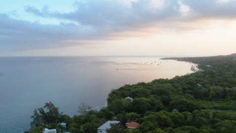 Ascending-Aerial-view-of-Caribbean-coast-with-boats,-turquoise-water,-clouds,-resorts-at-sunrise-with-island-background,-Roatan-island,west-end,-Honduras