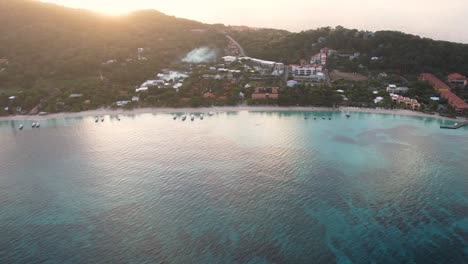 Cinematic-Aerial-view-of-Caribbean-coast-with-boats,-turquoise-water,-white-sand-beach,-resorts-during-sunrise,-island-background,-Roatan-island,west-end,-Honduras