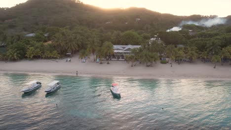 Cinematic-Aerial-view-of-Caribbean-coast-with-boats,-turquoise-water,-white-sand-beach,-resorts-at-sunrise-with-island-background,-Roatan-island,west-end,-Honduras