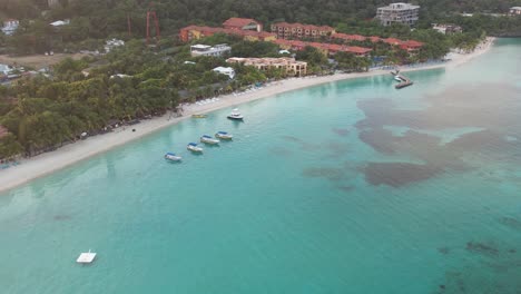 Cinematic-Aerial-view-of-Caribbean-reef-with-boats-in-turquoise-water,-beach,-resorts-at-sunrise,-island-background,-Roatan-island,-Honduras,-Central-America