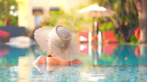 Back-view-Unrecognizable-woman-in-sunhat-resting-in-exotic-hotel-swimming-pool,-blured-lounge-deckchairs-and-umbrellas-on-background-in-Thailand-daytime