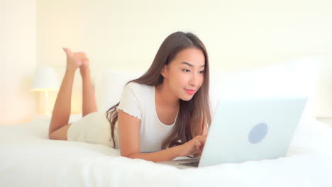 Beautiful-Exotic-Woman-Typing-on-Laptop-Keyboard,-Lying-on-Bed-in-Bright-Bedroom-Connecting-With-Friends,-Full-Frame
