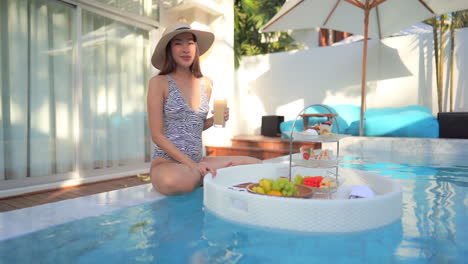 Sexy-Asian-Woman-Posing-to-Camera-at-Pool-With-Breakfast-on-Floating-Plate-on-Sunny-Morning-in-Luxury-Tropical-Hotel,-Full-Frame