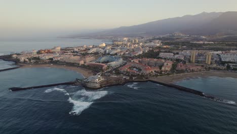 Playa-de-las-Americas-coastal-skyline,-Tenerife,-dawn-sunrise-aerial-drone-orbit