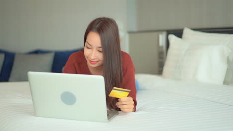 Hübsche-Asiatische-Frau,-Die-Online-Mit-Laptop-Und-Bankkreditkarte-Im-Schlafzimmer-Einkauft