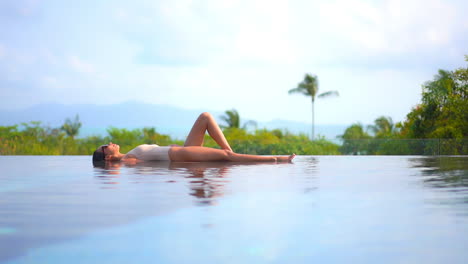 Slender-Woman-in-Swimsuit-Lies-on-Infinity-Pool-Border-in-Tropical-Landscape-on-Sunny-Day,-Full-Frame