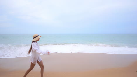 Slow-motion-of-a-cute,-young-woman-in-a-white-bathing-suit-walking-across-a-sandy-beach-as-the-waves-come-in
