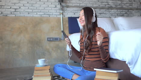 Mujer-Asiática-Feliz-Escuchando-Música-Desde-Un-Smartphone-Con-Auriculares-Y-Moviéndose-Al-Ritmo-De-La-Canción-Junto-A-La-Cama,-Fotograma-Completo-En-Cámara-Lenta
