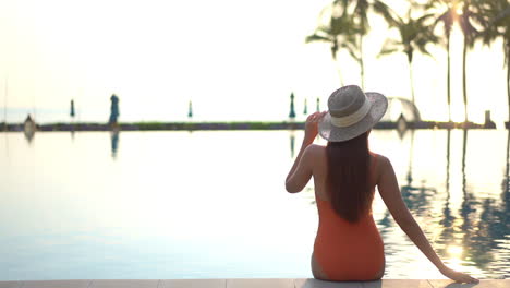 Back-view-of-woman-sitting-on-pool-edge-at-sunset-with-orange-swimsuit-and-holding-wide-hat