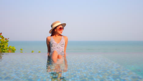 Young-woman-enjoying-rest-in-infinity-pool-outdoor-at-sunset-wearing-Sunhat,-red-sunglasses-and-spotted-monokini-style-swimwear