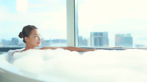 Pretty-Exotic-Woman-Relaxing-in-Bathtub-Full-of-Foam-With-Cityscape-View-Behind-Window,-Full-Frame