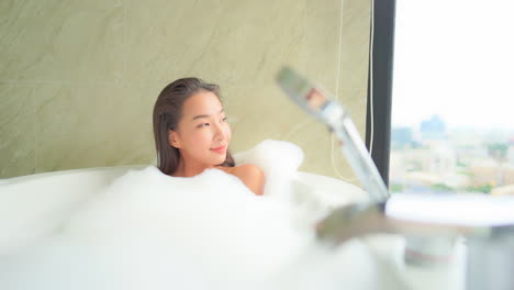 Schöne-Asiatische-Frau-In-Einer-Badewanne-Voller-Schaum,-Die-Durch-Das-Fenster-Schaut,-Spa-tag-entspannungsbehandlung,-Vollbild