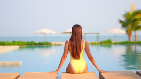 Rückansicht-Der-Frau,-Die-In-Gelben-Monokini,-Sonnenschirmen-Und-Palmen-Im-Hintergrund-Am-Rand-Des-Schwimmbads-In-Einem-Exotischen-Hotel-Auf-Bali-In-Strandnähe-Sitzt