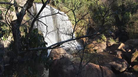 Luftpanoramaaufnahme-Von-Unten-Nach-Oben-Eines-Wunderschönen-Wasserfalls,-Der-Sich-Langsam-Hinter-Den-Bäumen-Offenbart