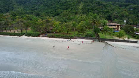 Aerial-orbiting-shot-of-kids-playing-on-the-beach-with-green-forest-with-mountains-in-the-background-and-beautiful-sea-with-gentle-waves-crashing