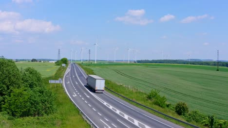 Large-White-Transport-Truck-driving-on-a-country-road-with-huge-ecological-Wind-Farm-in-background