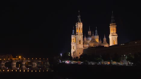 Traffic-Driving-Along-Ebre-River-Passing-By-Cathedral-Basilica-of-Our-Lady-of-the-Pillar-With-Puente-de-Piedra-At-Night-In-Zaragoza,-Spain