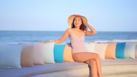 Sexy-Asiatisches-Model-Mit-Lila-Badeanzug-Und-Strohhut-Sitzt-Auf-Der-Strandsteinbank-Am-Meer-Und-Lächelt-Und-Entspannt-Sich-Auf-Gartenmöbeln-Im-Freien-Im-Thailand-Urlaub-Auf-Der-Terrasse-Des-Resorts