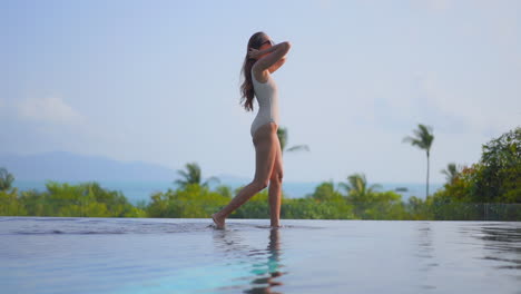 Petite-exotic-female-in-swimsuit-walking-on-water-of-infinity-pool-with-tropical-vegetation-in-background,-slow-motion