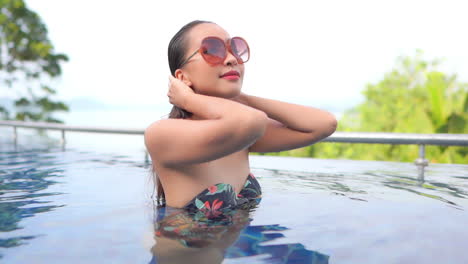 Young-brunette-Asian-woman-standing-inside-swimming-pool-wearing-a-flowery-bikini,-touching-her-wet-hair-rising-her-face-up-with-delight,-and-opening-her-eyes-in-Bali,-slow-motion