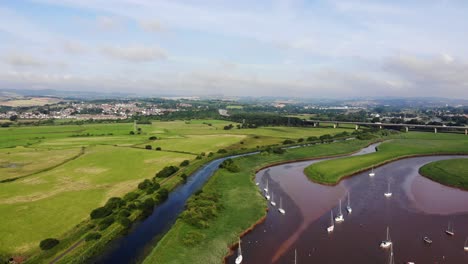 Aerial-Over-Sailboats-Anchored-In-River-Exe-In-Topsham,-Pan-Left-To-Green-Fields