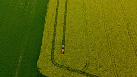 Farm-Tractor-with-spray-booms-spraying-pesticides-over-yellow-blooming-rapeseed-field,-aerial-pull-back