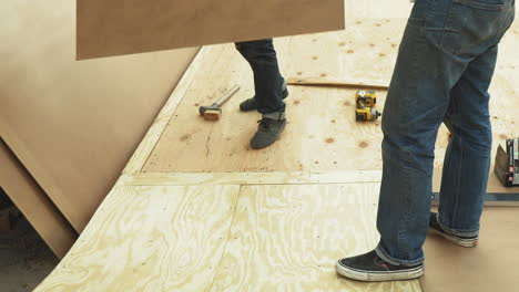 Zwei-Männer-In-Jeans-Heben-In-Der-Werkstatt-Sperrholz-Auf,-Um-Eine-DIY-Skateboardrampe-Zu-Bauen,-Kein-Gesicht,-Nur-Beine,-Unterkörper