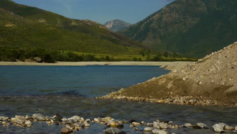 Creek-water-streaming-into-river-of-Vjosa-with-mountains-background-in-Albania