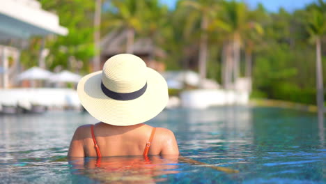Backside-view-of-an-Unrecognizable-Pretty-Young-Model-in-Swimming-Suit-and-Sunhat-Standing-Inside-the-Pool-of-Tropical-Island-Resort-in-Bali,-Indonesia,-blurred-palm-trees-on-background