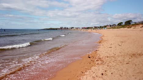 Golden-sands-and-gentle-waves-looking-towards-Earlsferry-on-the-east-coast-of-Fife-Scotland-with-beach-huts-and-paddle-boats-and-people-enjoying-the-water