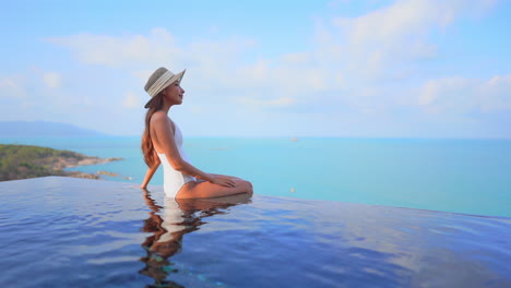 A-fit-young-healthy-woman-sits-on-the-edge-of-a-resort-pool-while-looking-out-over-the-ocean-horizon