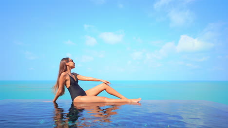 Sexy-exotic-female-in-swimsuit-sitting-on-infinity-pool-edge-enoying-in-stunning-view-of-tropical-sea-horizon,-slow-motion