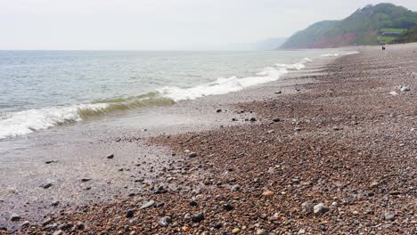 Small-waves-roll-onto-the-pebble-beach-of-Branscombe-in-Devon-on-the-English-Channel-coast-on-a-warm-hazy-day