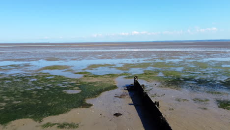Low-drone-flight-over-an-old-wooden-groyne-and-out-over-mudflats-of-a-tidal-estuary-on-a-bright-and-sunny-afternoon-with-a-bright-blue-sky
