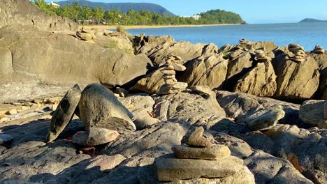 smooth-rising-shot-over-a-rocky-shoreline-with-many-rock-cairns-piled-up-among-the-rocks,-as-the-view-opens-out-to-a-broad-tropical-and-sunlit-bay-in-the-background