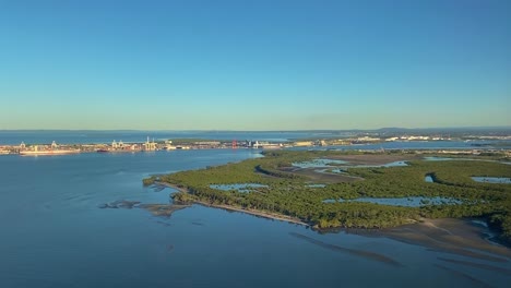 Low-aerial-shot-over-mangroves-and-marshlands-with-port-infrastructure,-waterways-and-coastal-pools,-with-a-view-to-the-horizon-under-a-brilliant-blue-sky