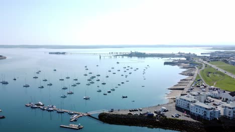 Sunny-Holyhead-harbour-breakwater-maritime-yachts-docked-along-transparent-calm-blue-shoreline-aerial-descending-view