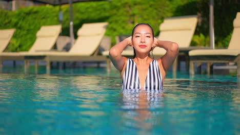 Feminine-Asian-woman-squeeze-water-out-of-hair-while-bathing-in-a-swimming-pool,-she-is-in-water-up-to-the-neck-and-touching-her-wet-hair,-face-close-up,-Dominican-Republic